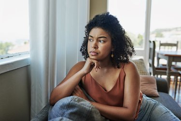 an adult sits in a chair at home looking worried with their chin resting on their hand looking out the window