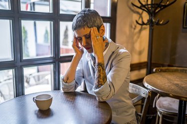 Person with a short hair cut and tattoo sleeves sitting at a wooden table holding their head due to pain from stroke mimics