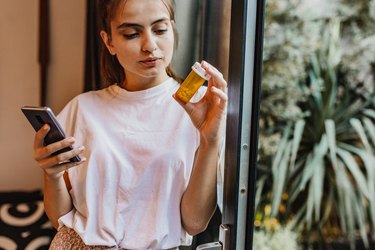 Person with brown hair in a ponytail, wearing a pink t-shirt, holding their phone in one hand and methotrexate pill bottle in another hand