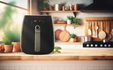A black air fryer with a silver handle placed on a wooden kitchen counter surrounded by kitchen supplies and utensils